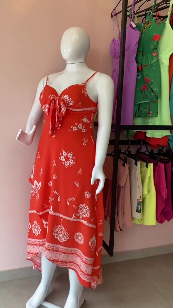 Manequim com um vestido vermelho estampado e uma arara com roupas femininas coloridas ao fundo, na parede rosada da boutique