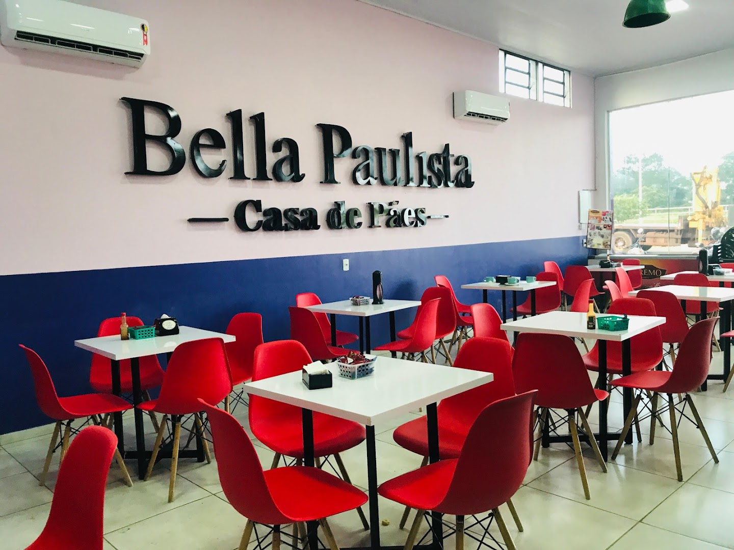 Salão interno da padaria com mesas brancas e cadeiras vermelhas