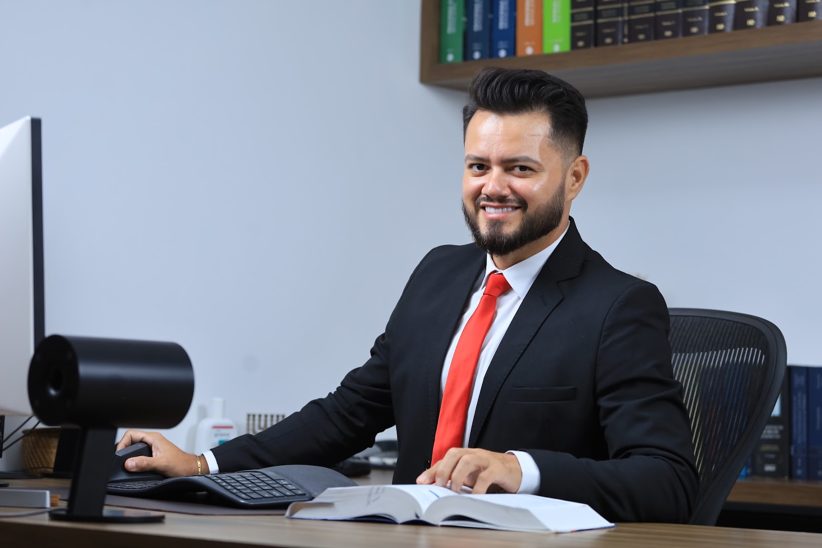 Dr. José Alex da Silva Bernardes em sua mesa de trabalho, de terno e gravata vermelha