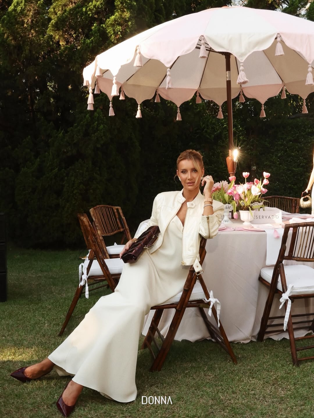 Modelo em vestido longo off white e jaqueta, sentada sob guarda sol em mesa posta