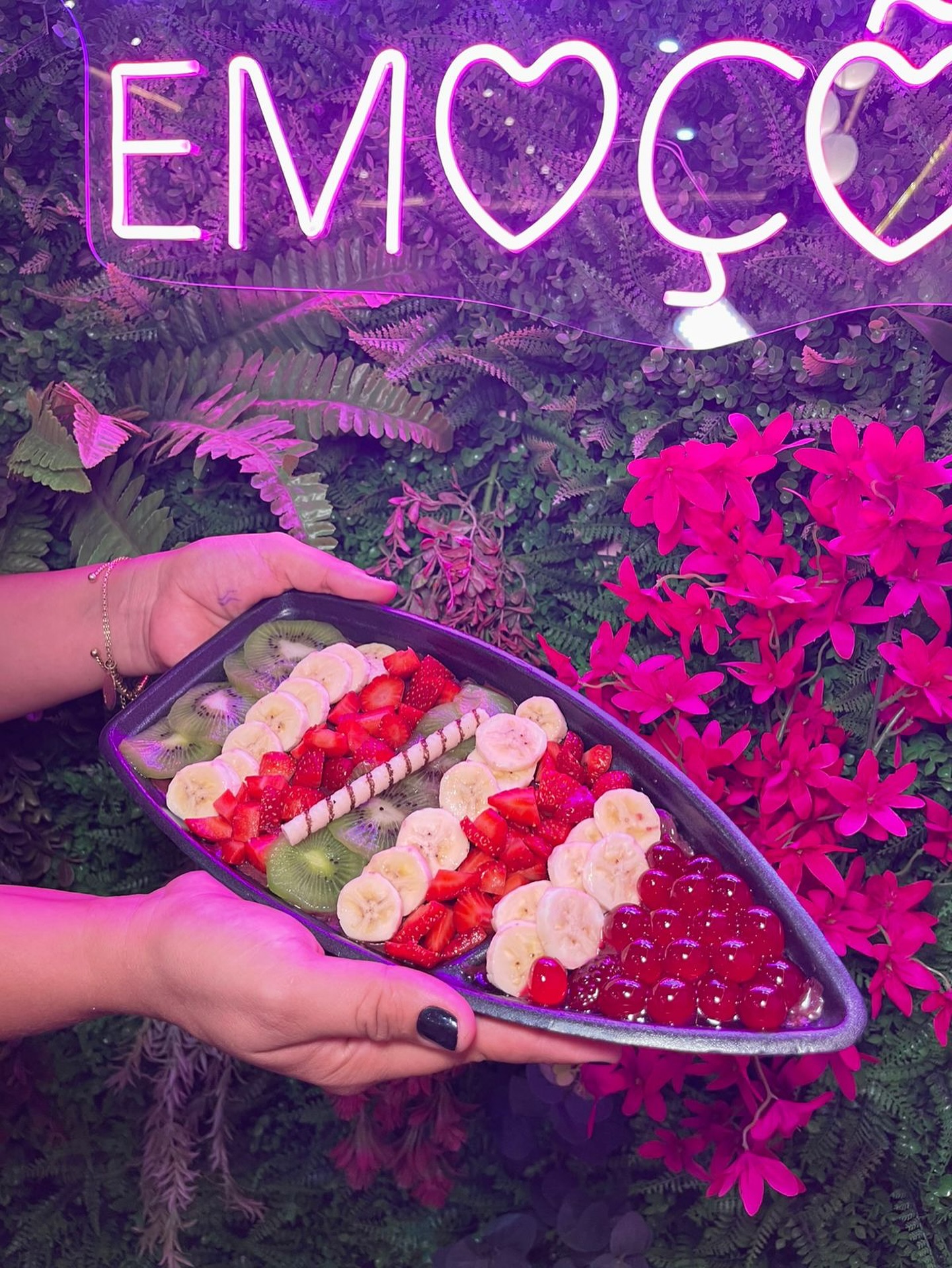 Tábua de frutas com morango, banana e kiwi servida sob o letreiro de neon da Geerrê