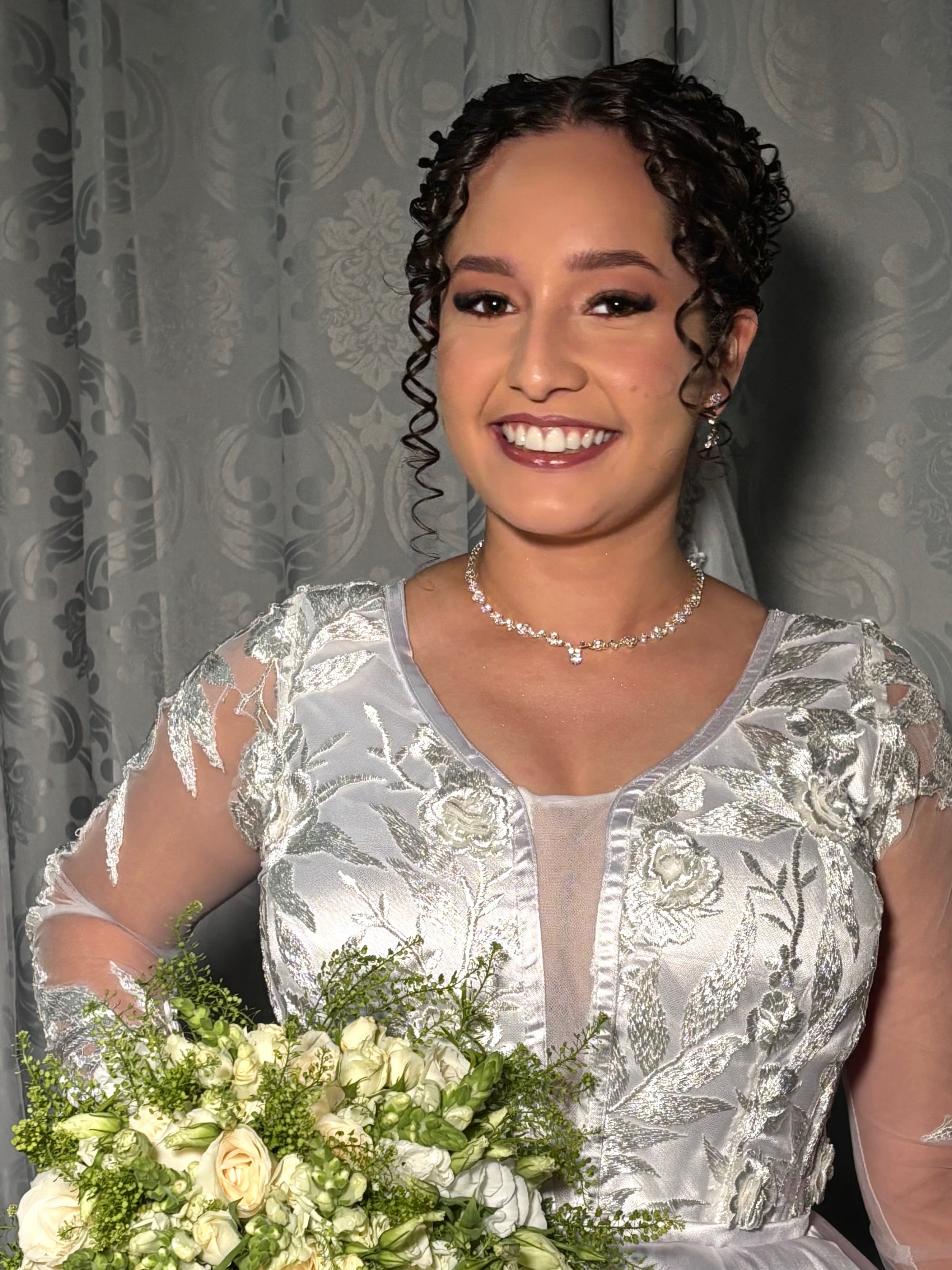 Noiva com maquiagem e penteado por Luzi Makeup, sorrindo com buquê de flores