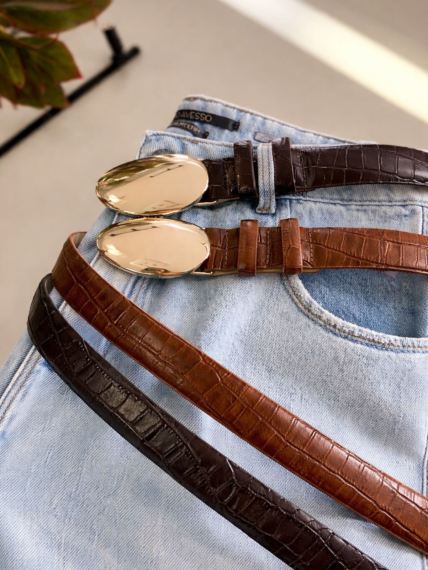 Cintos de couro marrom e preto com fivela oval dourada sobre calça jeans