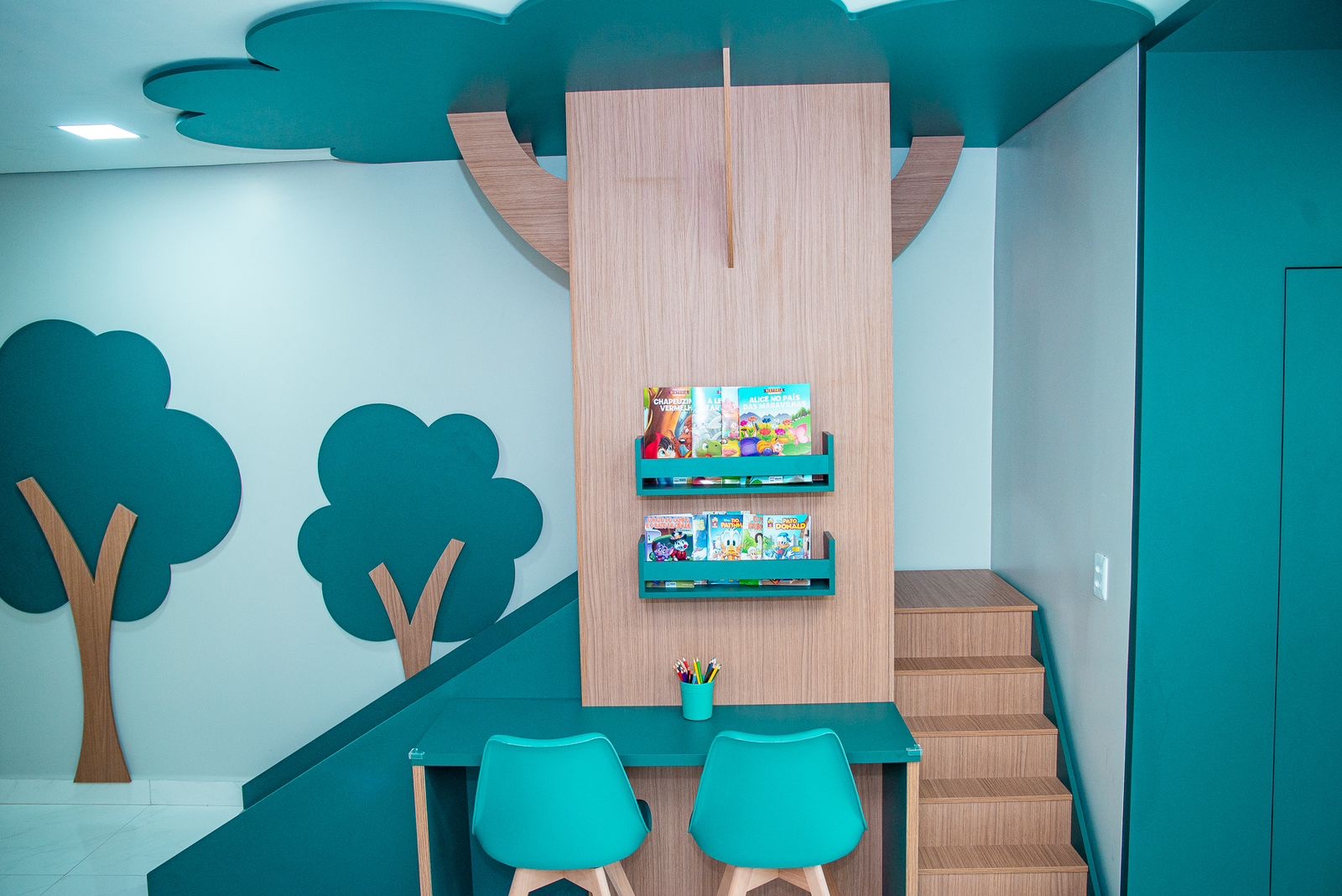 Cantinho infantil com estante em formato de árvore, cadeirinhas e livros