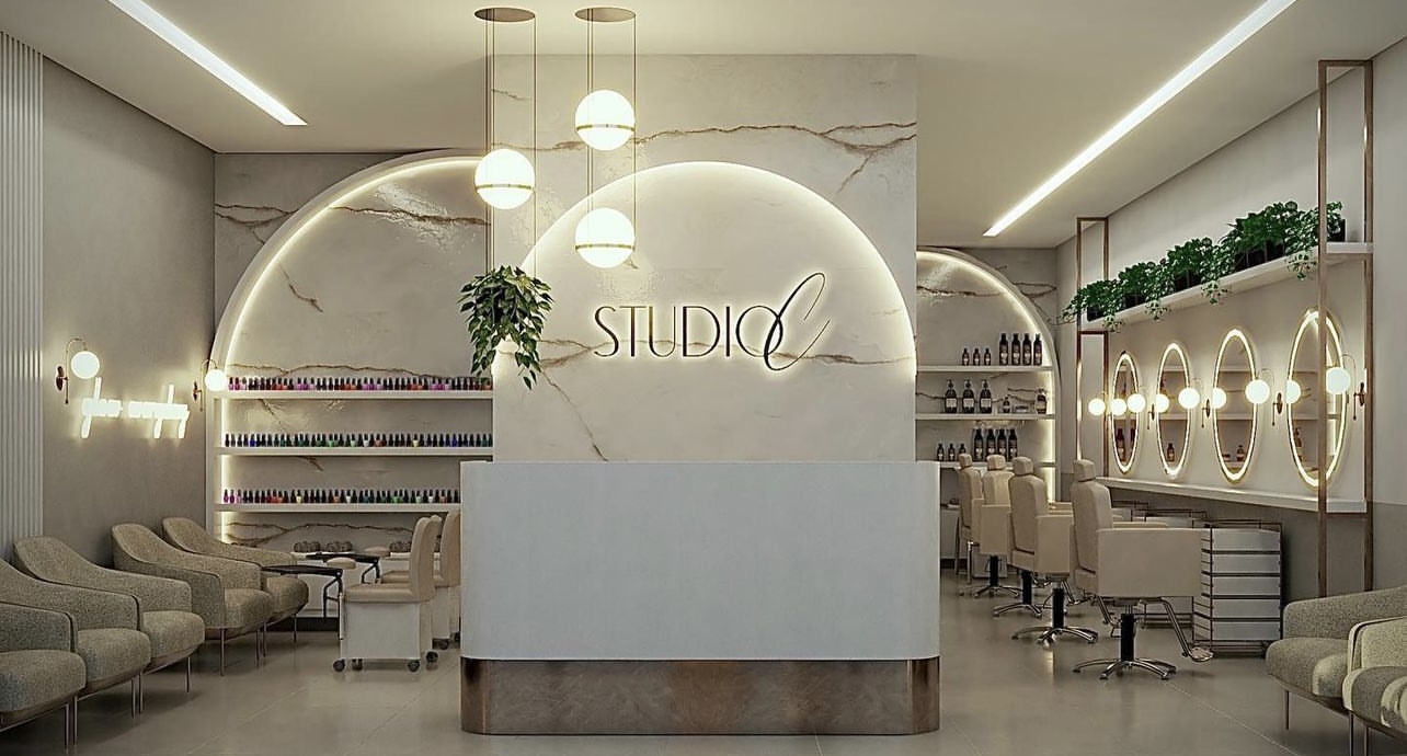 Interior do Studio C Exclusive Beauty, com paredes de mármore claro, arco iluminado com o logo do salão, luminárias globo douradas e poltronas em tom nude.