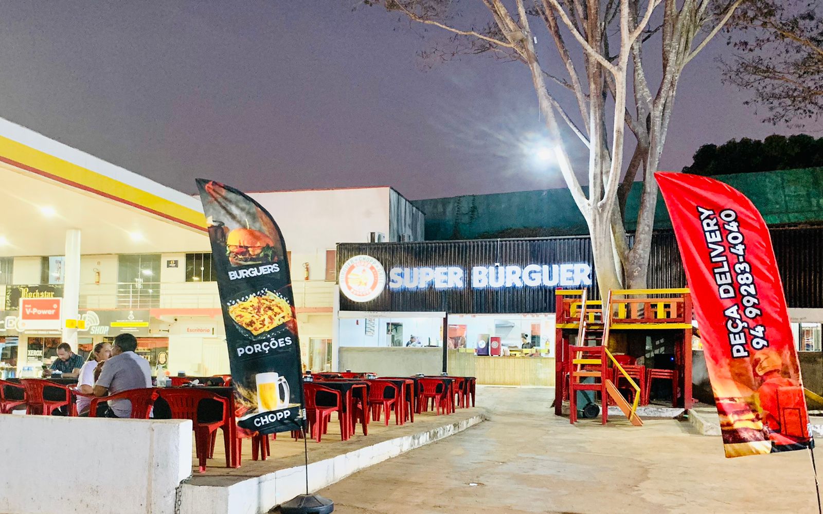 Fachada da Super Burguer à noite com mesas na frente